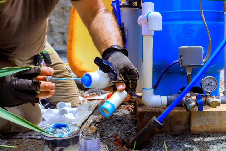 Técnico de luvas montando conexões de PVC ao lado do reservatório azul de pressurização, em assistência técnica de poço artesiano no Rio de Janeiro