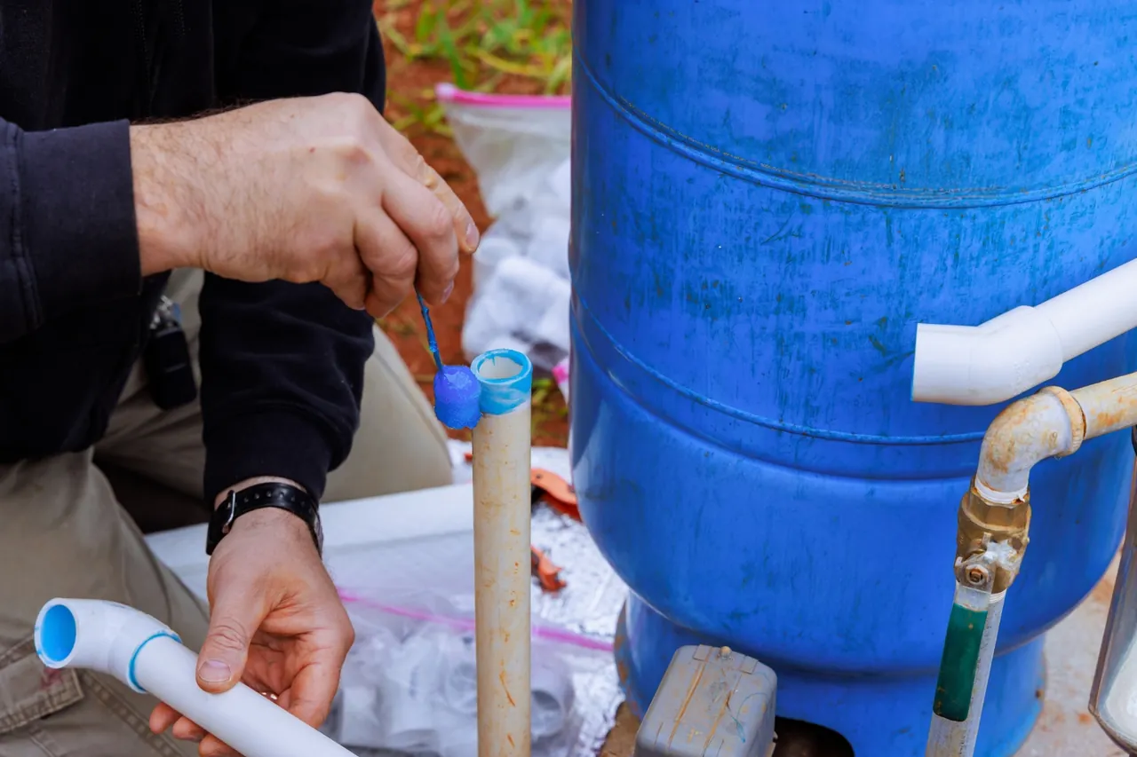 Mão de técnico com luva colocando tampa em tubo de PVC próximo ao tanque azul na instalação da bomba